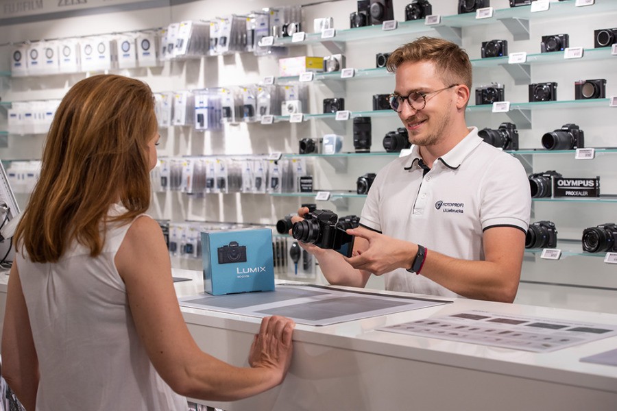Verkäufer präsentiert Kamera einer Kundin in einem Elektronikgeschäft mit Wand voller Kameraprodukte.